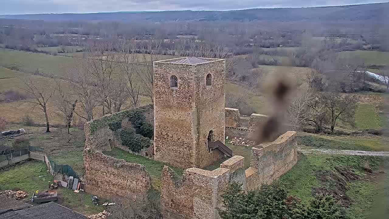 An overhead view shows the ruins of a stone building, including a relatively intact square tower, situated in a rural landscape under an overcast sky.