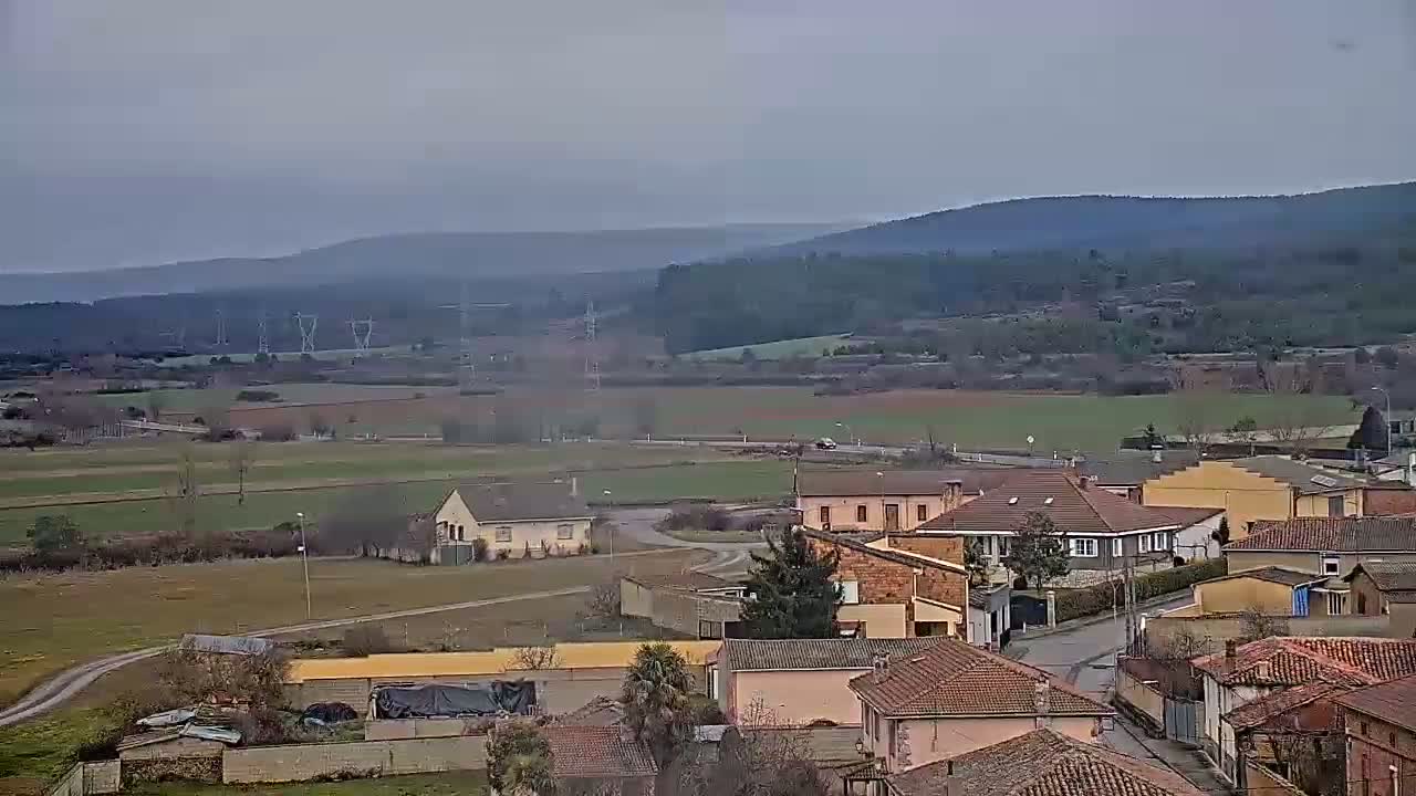 A small village of low-slung buildings sits in a valley under an overcast sky, with rolling hills and distant mountains in the background.