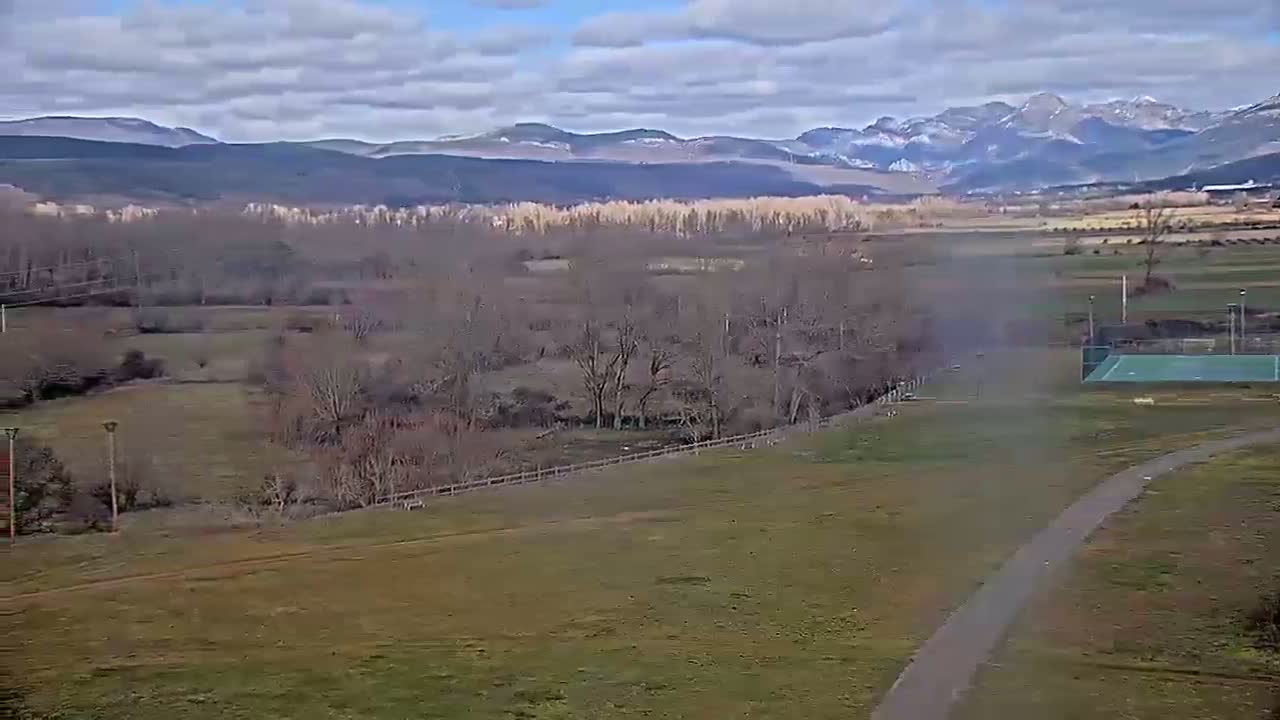 A partly cloudy day reveals a wide, grassy field with a paved path, a tennis court, and a background of bare trees, a river, and snow-capped mountains in the distance.