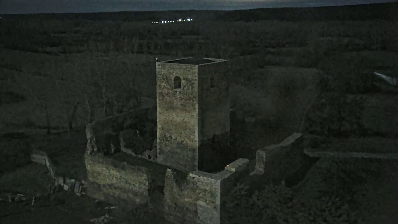 An aerial view at night shows the stone ruins of a tower and surrounding walls, set against a dark landscape under a dimly lit sky.