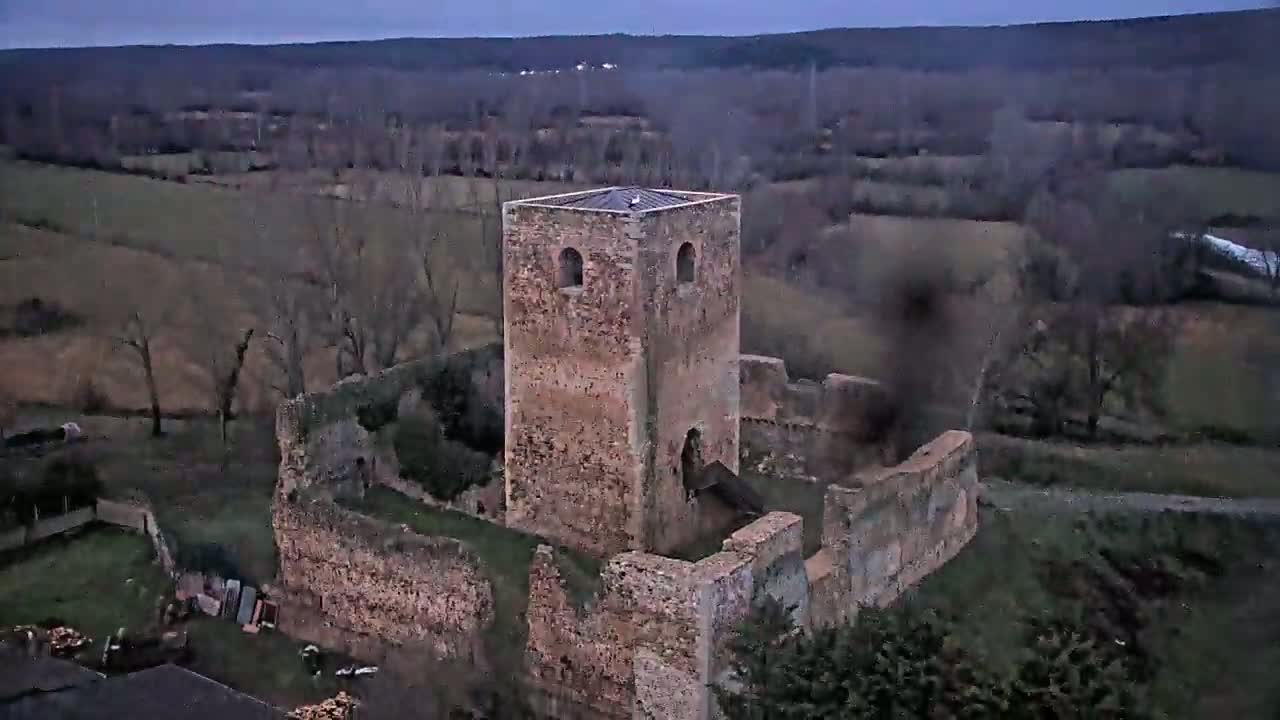 An aerial view shows the ruins of a stone tower and surrounding walls, situated in a rural landscape under a cloudy sky.