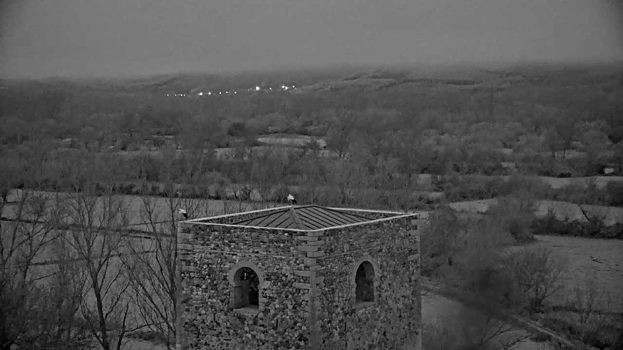 A stone structure with a pitched roof sits atop a hill overlooking a valley of bare trees and fields under an overcast sky.