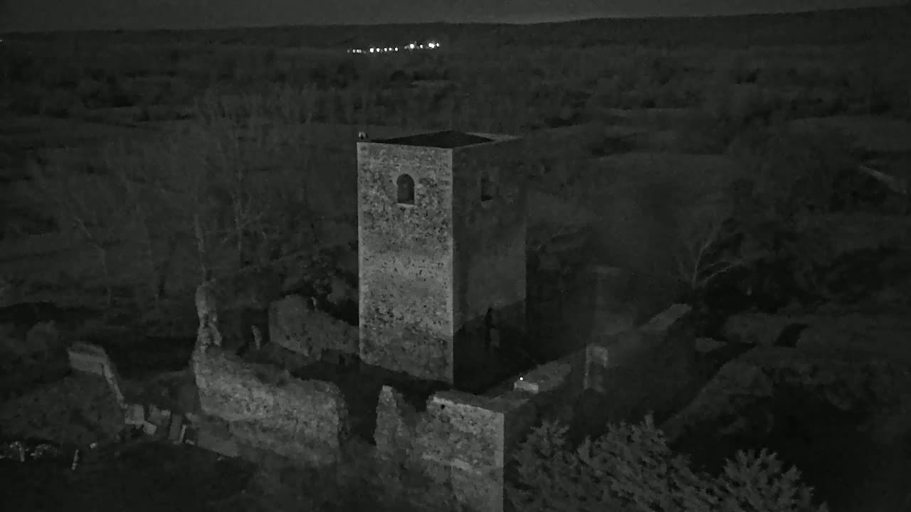 An aerial nighttime view shows the stone ruins of a tower and surrounding walls, situated in a landscape of dark trees under a clear, night sky.