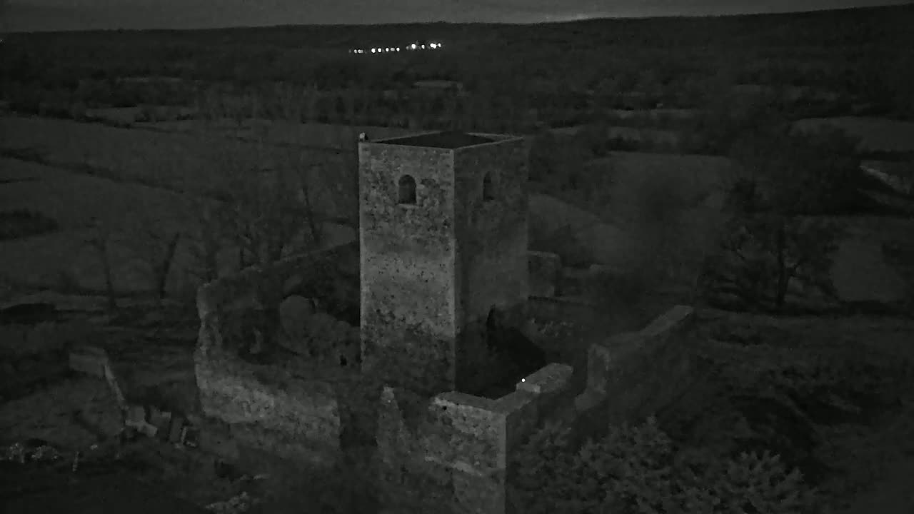 An aerial nighttime view shows the stone ruins of a tower and surrounding buildings in a dark, rural landscape.