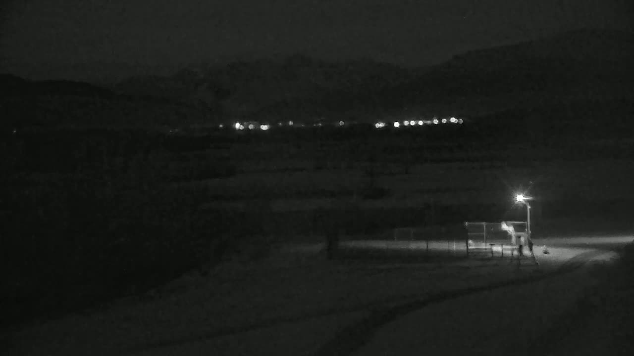 A snow-covered landscape at night, illuminated by a single lamppost near a fence, shows distant lights of a town against a dark mountain range.