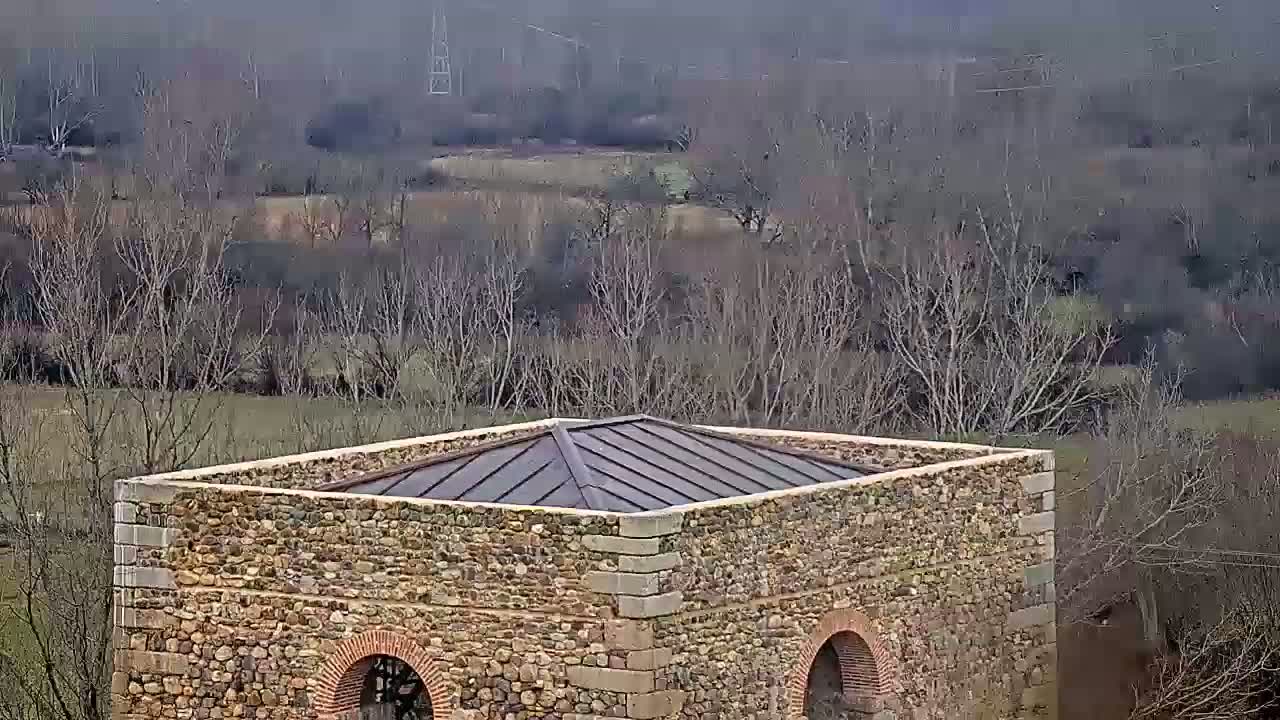 A stone structure with a dark metal roof sits atop a hill overlooking a valley of leafless trees under an overcast sky.