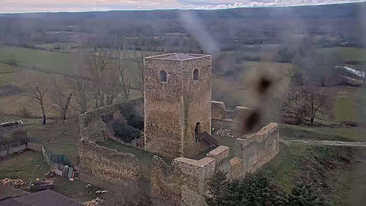 An aerial view shows the stone ruins of a castle, including a tall standing tower, on a partly cloudy day in a rural setting.