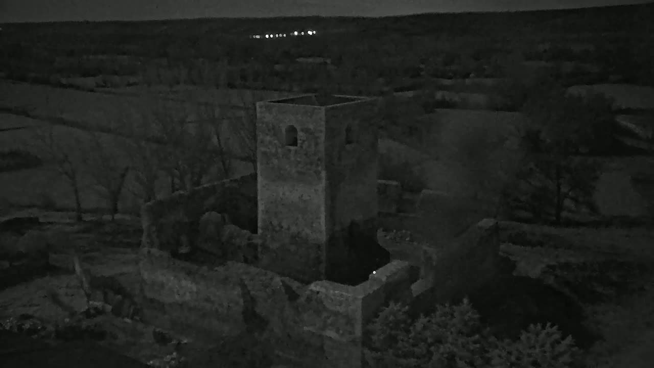 An aerial nighttime view shows the ruins of a stone structure, possibly a tower or small castle, surrounded by dark trees under a dark sky.