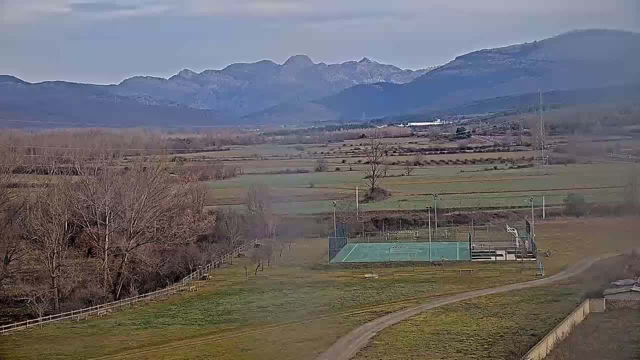 An outdoor basketball court sits in a grassy field, overlooked by bare winter trees and distant mountains under a cloudy sky.