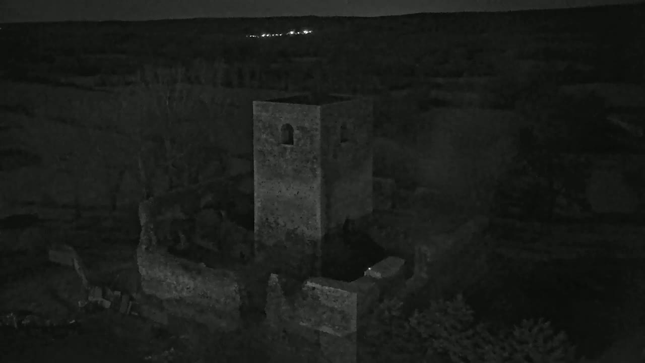 An aerial nighttime view shows the stone ruins of a tower and surrounding walls under a dark sky with a few distant lights visible on the horizon.