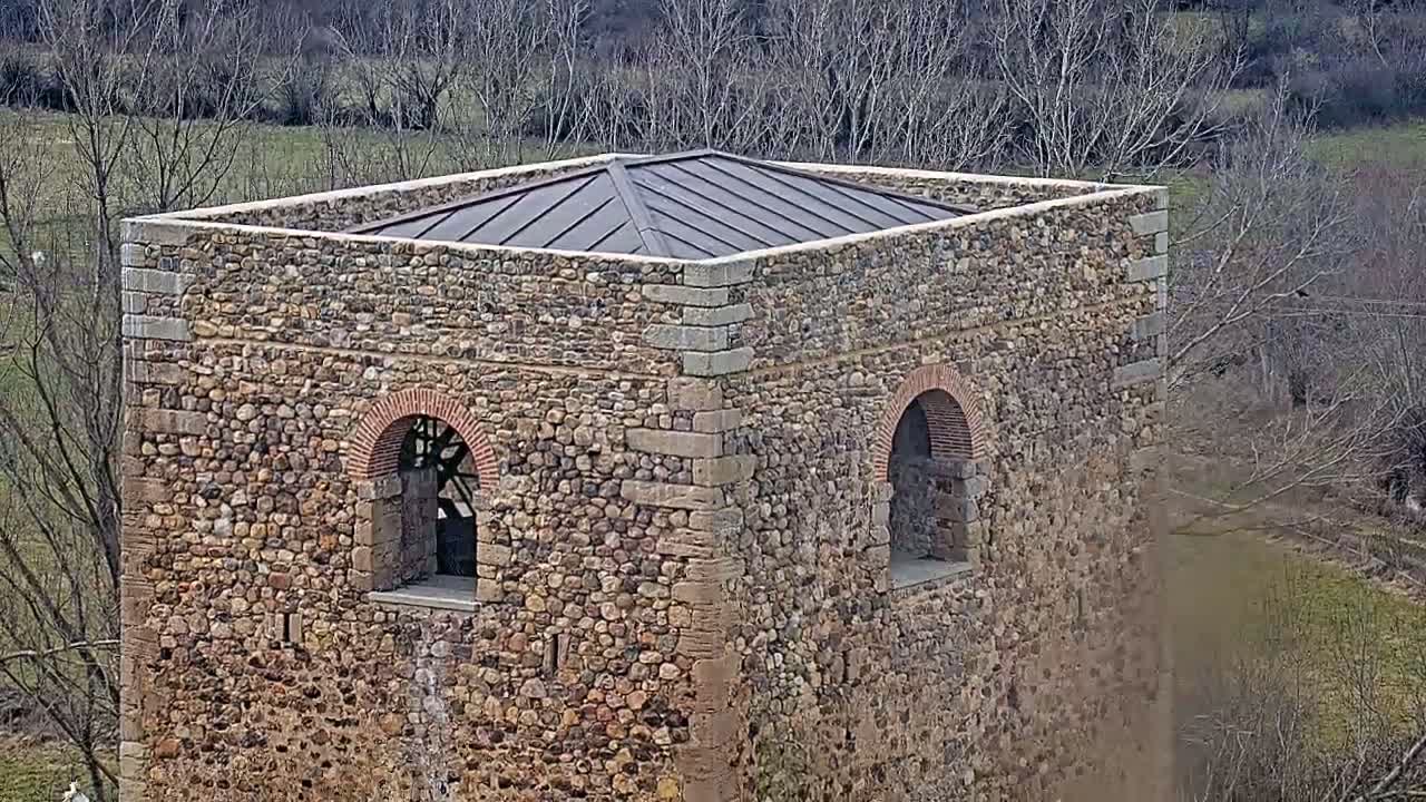A stone structure with a flat, dark metal roof, featuring two arched windows, is situated in a landscape of bare trees and a grassy field.