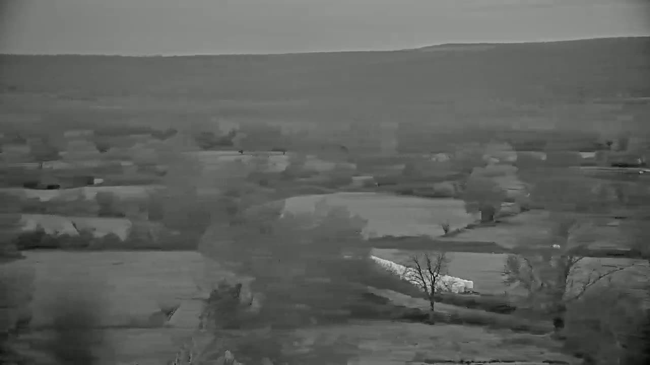 A grayscale aerial view shows a rural landscape with fields, trees, and a distant hill under overcast skies.