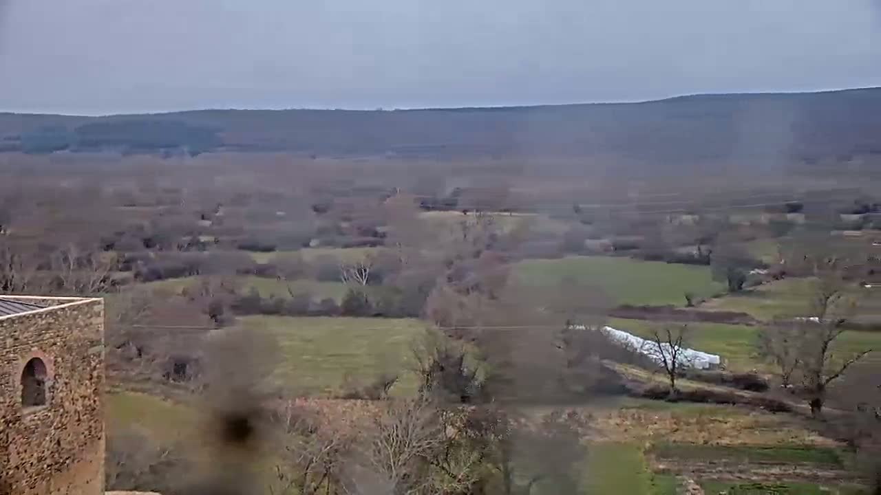 A sprawling, overcast landscape of fields, trees, and a stone structure, viewed from a high vantage point.