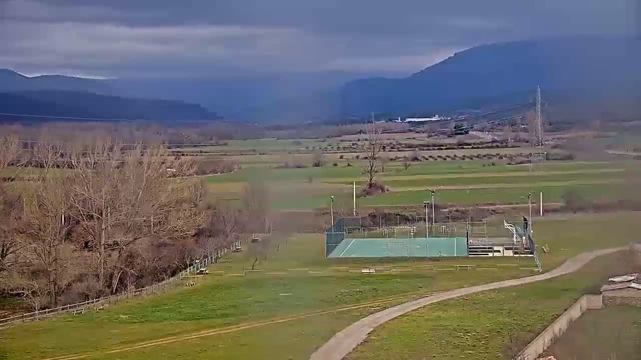 An outdoor sports court sits in a grassy field, overlooked by bare trees and distant mountains under a cloudy sky.