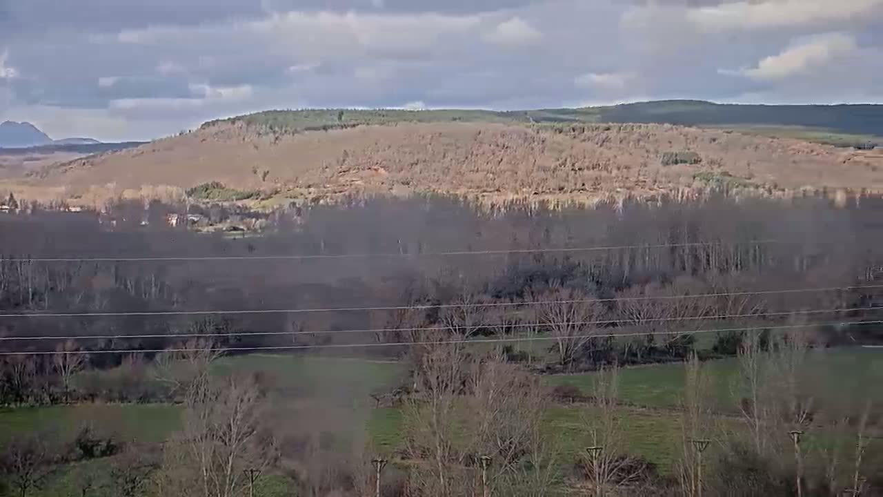 A hazy landscape under a partly cloudy sky shows a valley of bare winter trees and fields, beyond which is a hillside covered in scrubby vegetation.