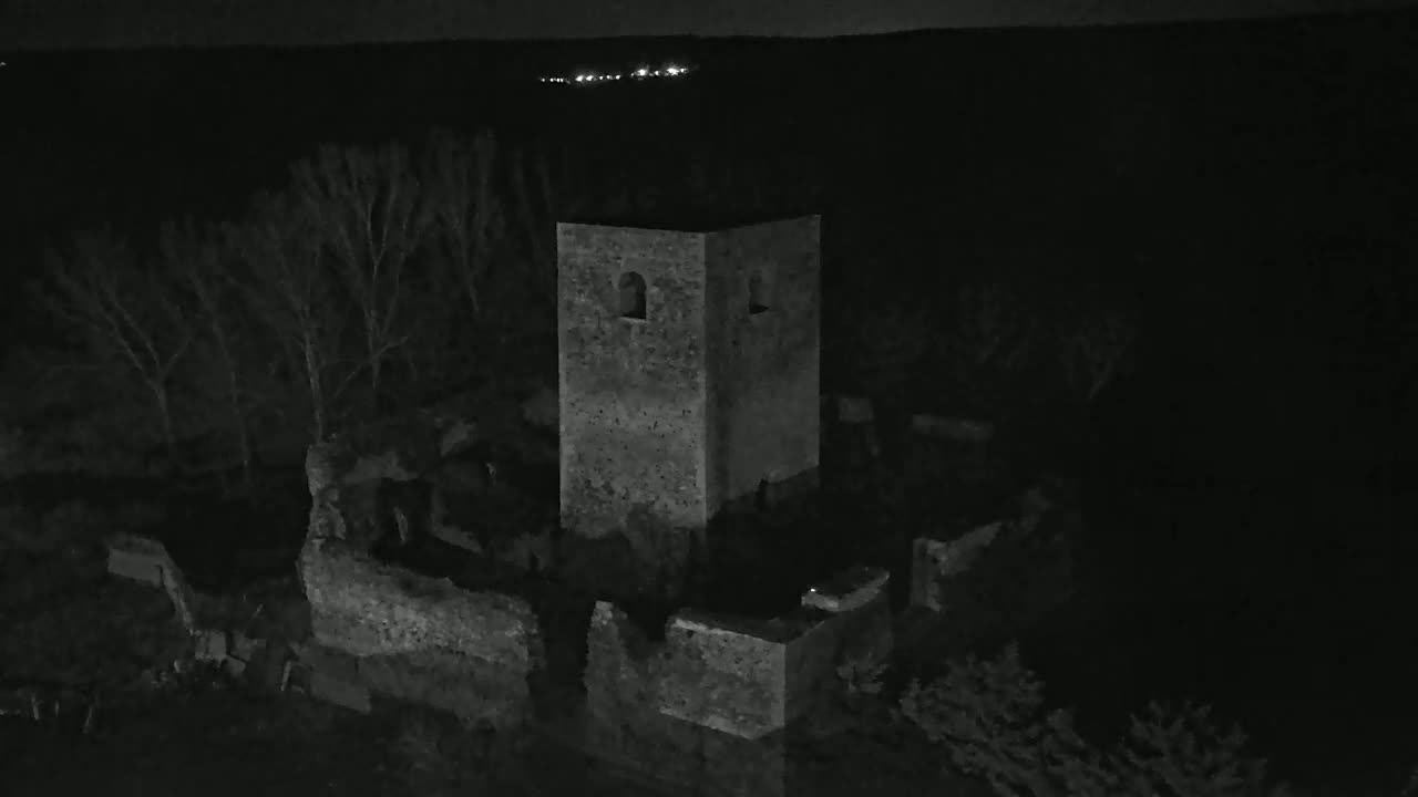 An aerial view at night shows the stone ruins of a building, including a standing tower, surrounded by bare trees under a dark sky.