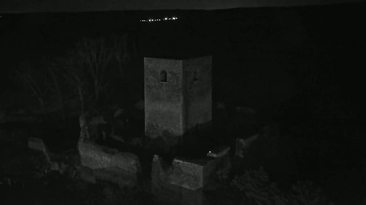 A stone tower stands at night amidst dark, indistinct structures, with a distant line of lights visible on the horizon.