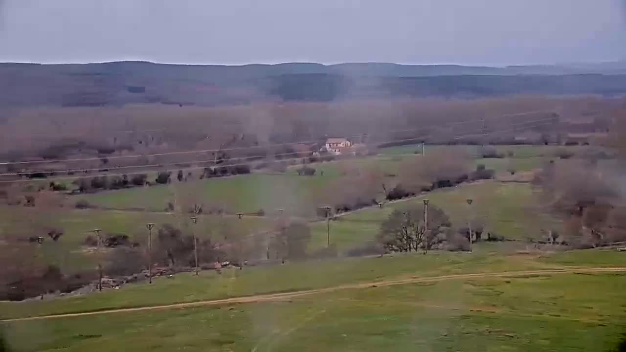 A hazy, overcast day reveals a sprawling landscape of rolling green hills, dotted with sparse trees, a lone farmhouse, and power lines.