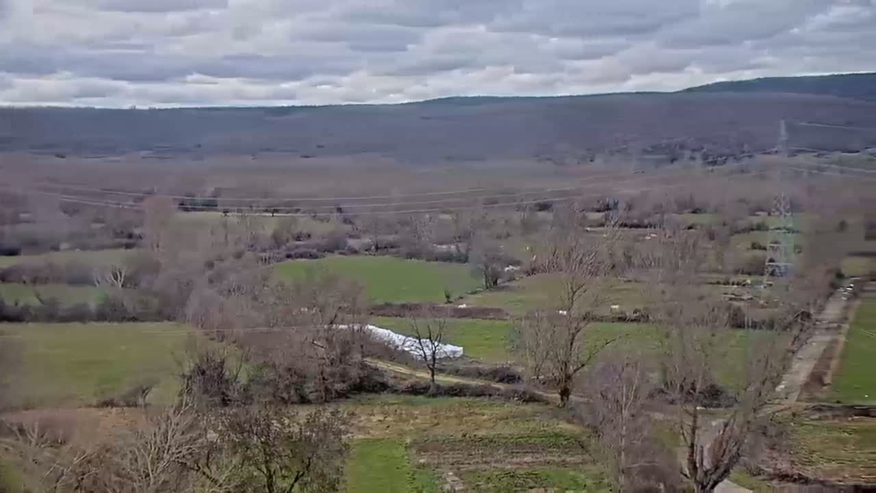A high-angle view of a rural landscape under a cloudy sky shows fields, leafless trees, and a single white structure in a valley, with hills in the background.