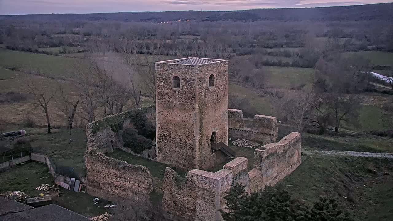 Stone ruins of a tower and walls stand in a rural landscape under a dusky sky.