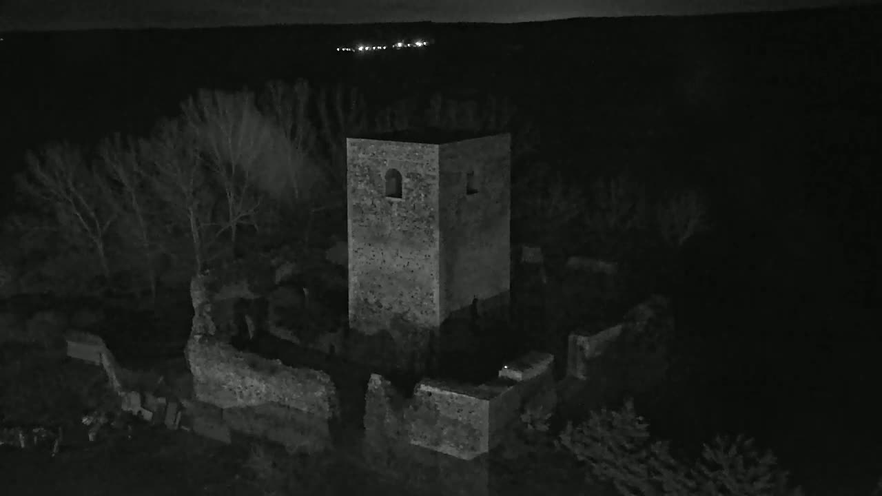 An aerial nighttime view shows the stone ruins of a tower and surrounding walls, nestled among bare trees under a dark sky.