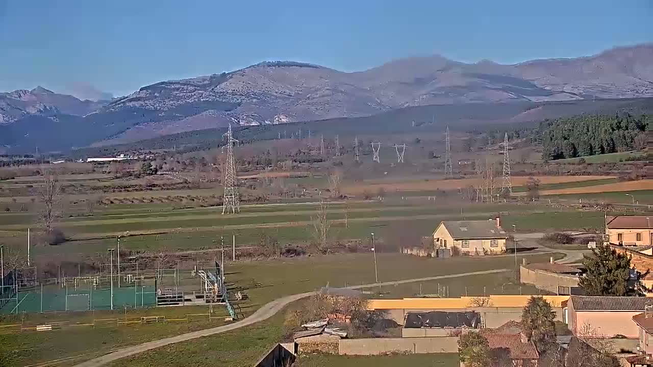 A panoramic view shows a rural landscape under a clear, sunny sky, with a small village nestled among fields and power lines, and snow-capped mountains in the background.
