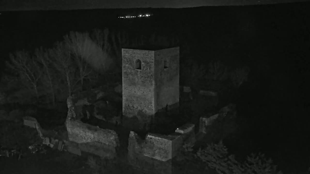 An aerial nighttime view shows stone ruins, including a standing tower, surrounded by dark trees under a clear, dark sky.