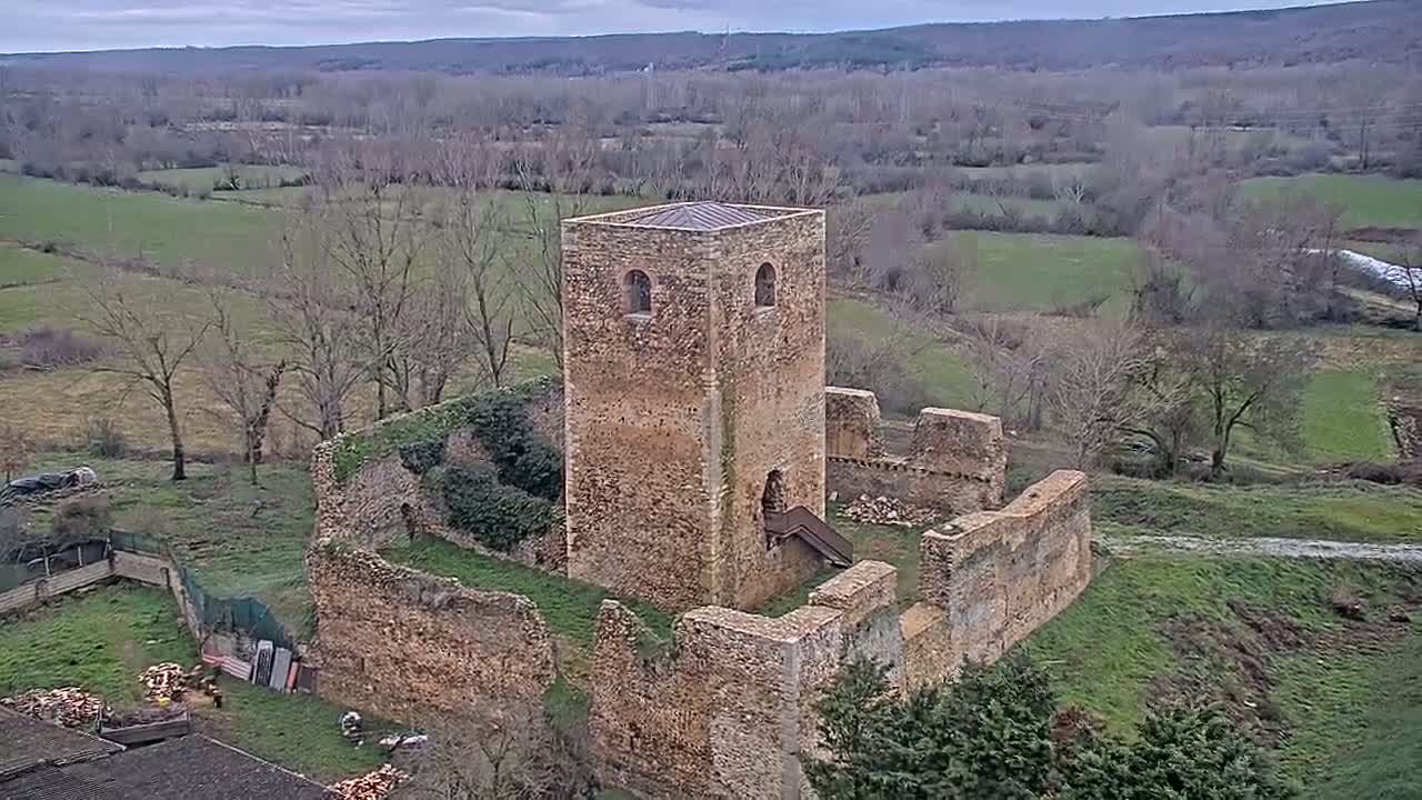 An aerial view shows the ruins of a stone tower and surrounding walls, situated in a verdant, hilly landscape under an overcast sky.