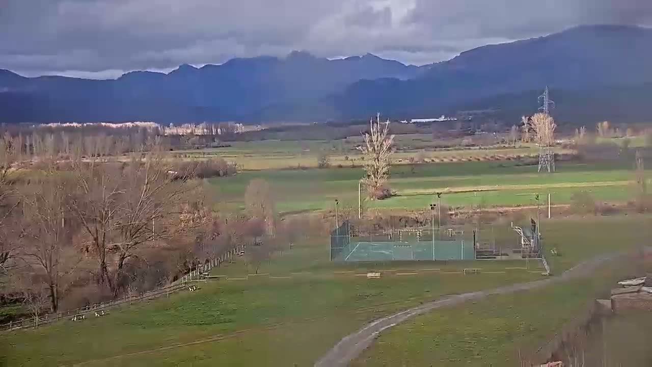 A view of a rural landscape under a partly cloudy sky shows a small sports court, bare winter trees, and distant mountains.