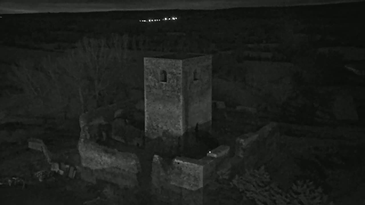 A stone tower, part of a larger ruin, stands at night under a dark sky with distant lights visible on the horizon.
