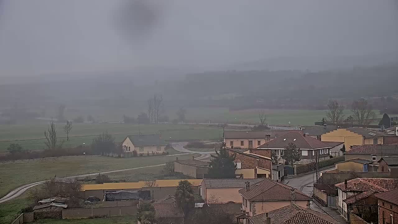 A foggy, overcast view of a small town nestled in a valley, with houses and buildings clustered together, and surrounding fields stretching out to the horizon.