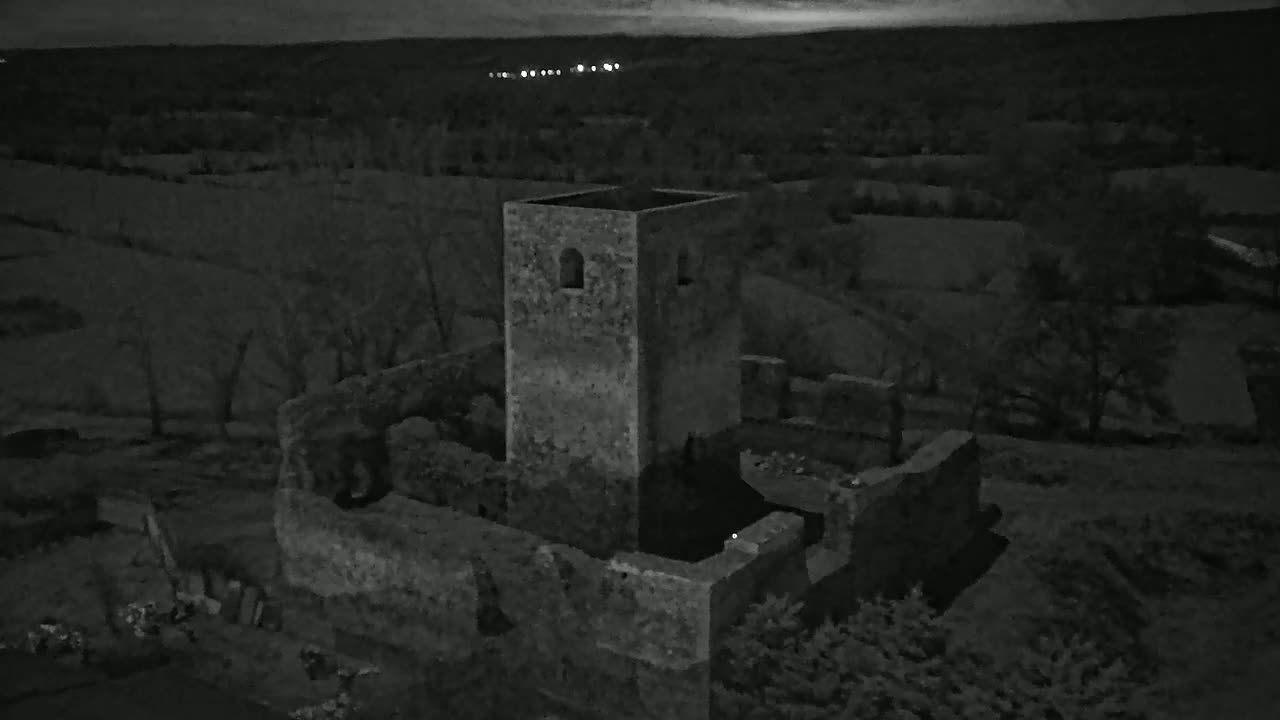 An aerial view at night shows the ruins of a stone tower and surrounding structures, set against a dark landscape under a mostly clear sky.