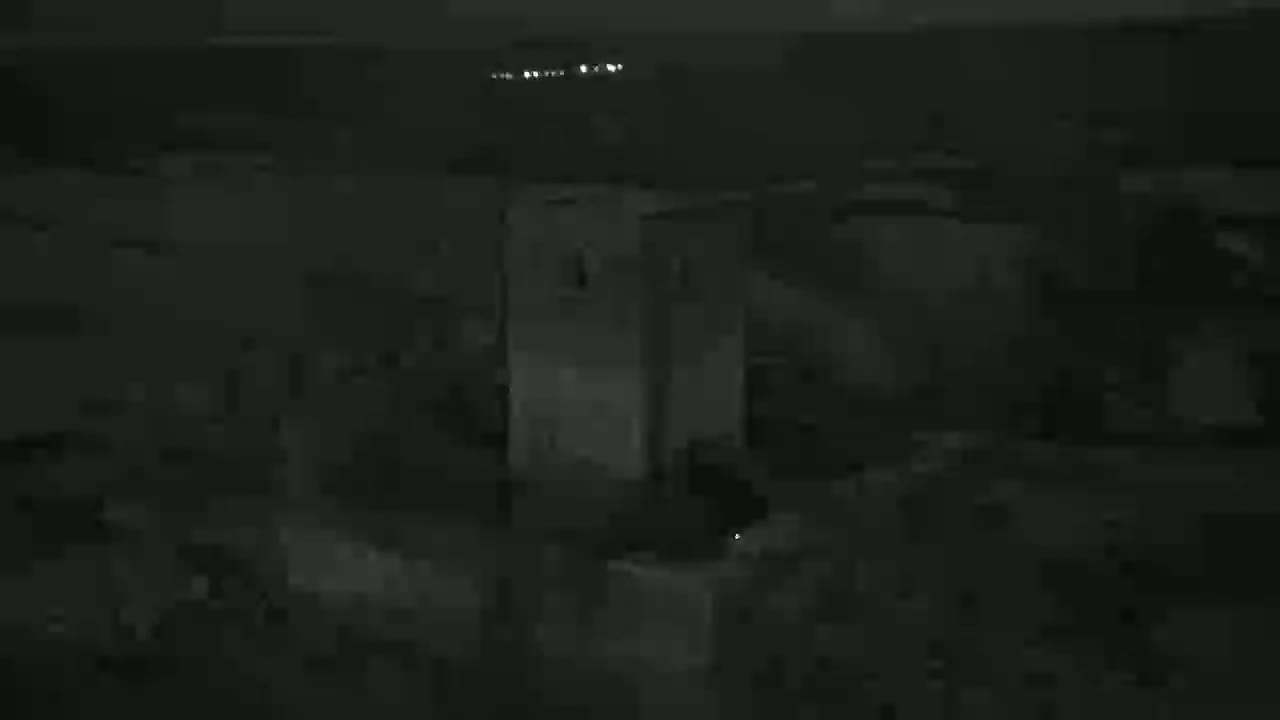 A dark, nighttime image shows a tall, light-colored structure, possibly a tower or building, surrounded by darker, indistinct shapes in a dimly lit area.