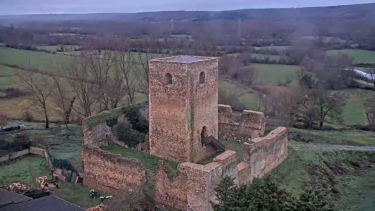 A stone tower and the ruins of a larger structure stand on a grassy hill overlooking a valley on an overcast day.