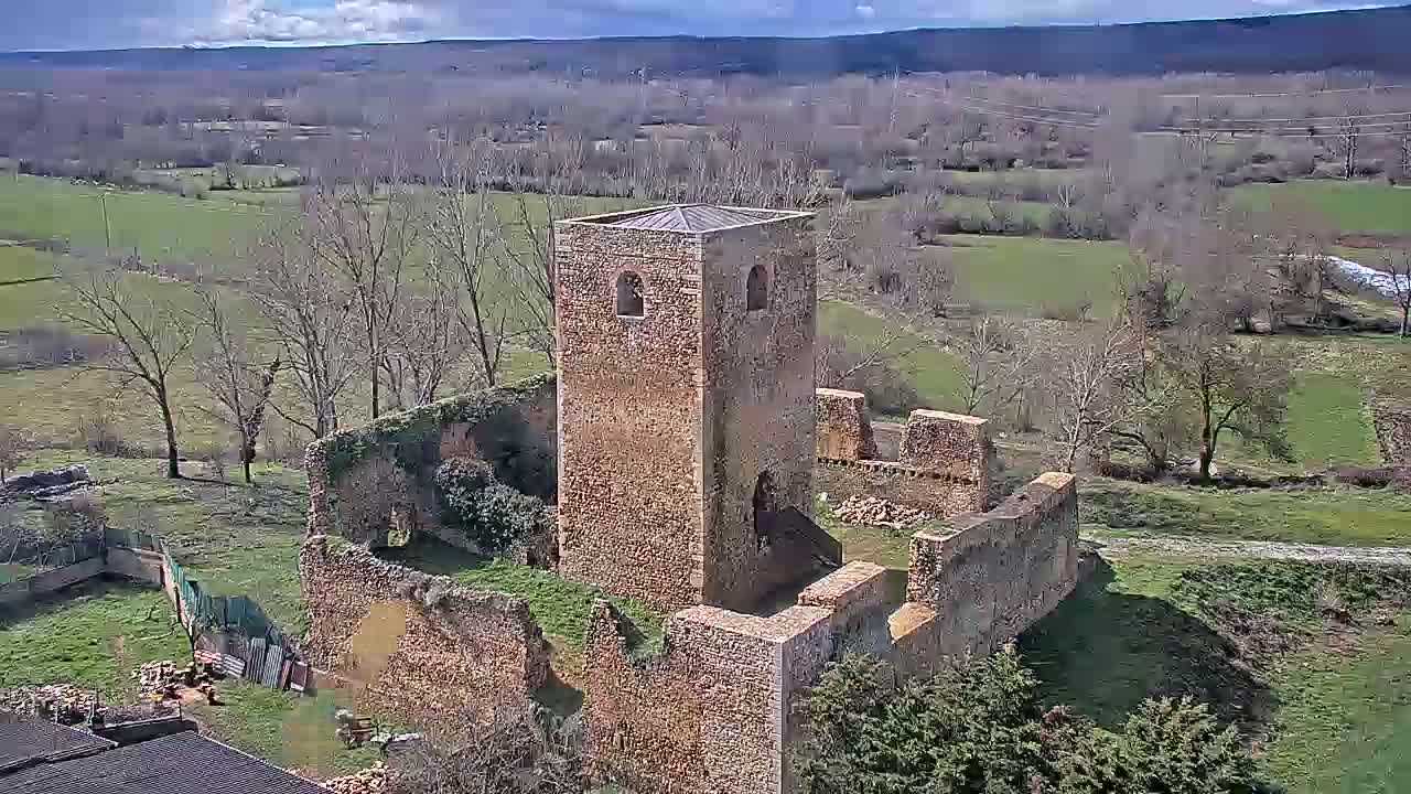 An aerial view shows the stone ruins of a building, possibly a church or castle, on a partly sunny day with a background of rolling hills and sparse trees.