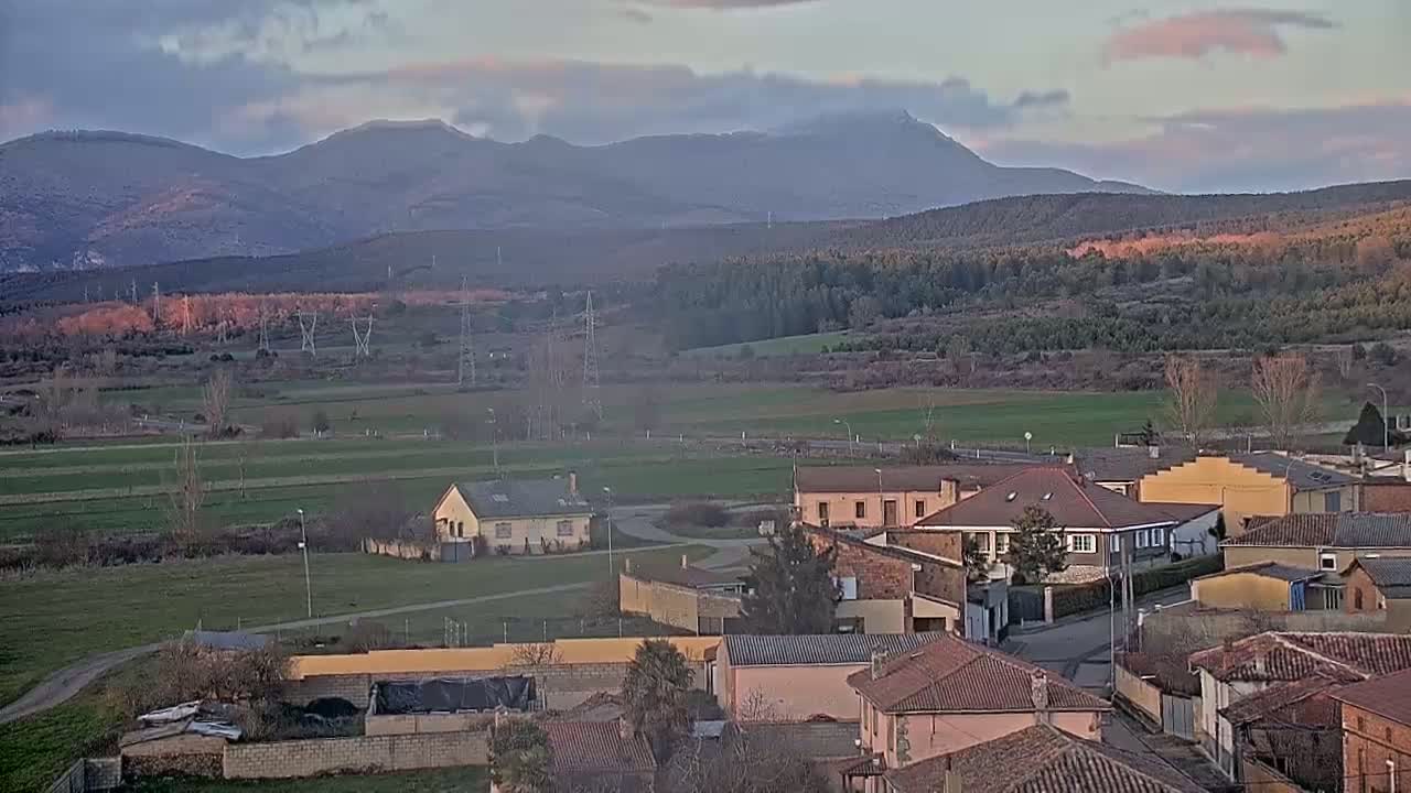 A small town with red-tiled roofs sits in a valley beneath a range of mountains under a mostly cloudy sky.