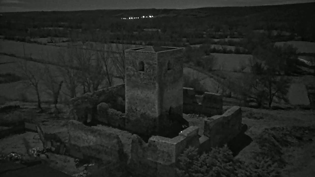 An aerial nighttime view shows the stone ruins of a tower and surrounding buildings amidst bare winter trees and fields under a dark sky.