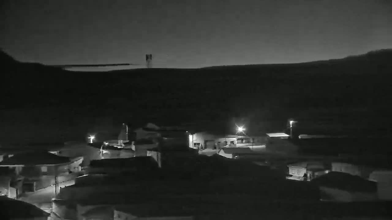 A nighttime, low-light view of a small village nestled against a dark hill under a clear, dark sky.