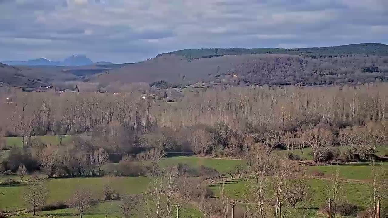 A view of a valley with bare trees and green fields under a partly cloudy sky, with distant mountains in the background.