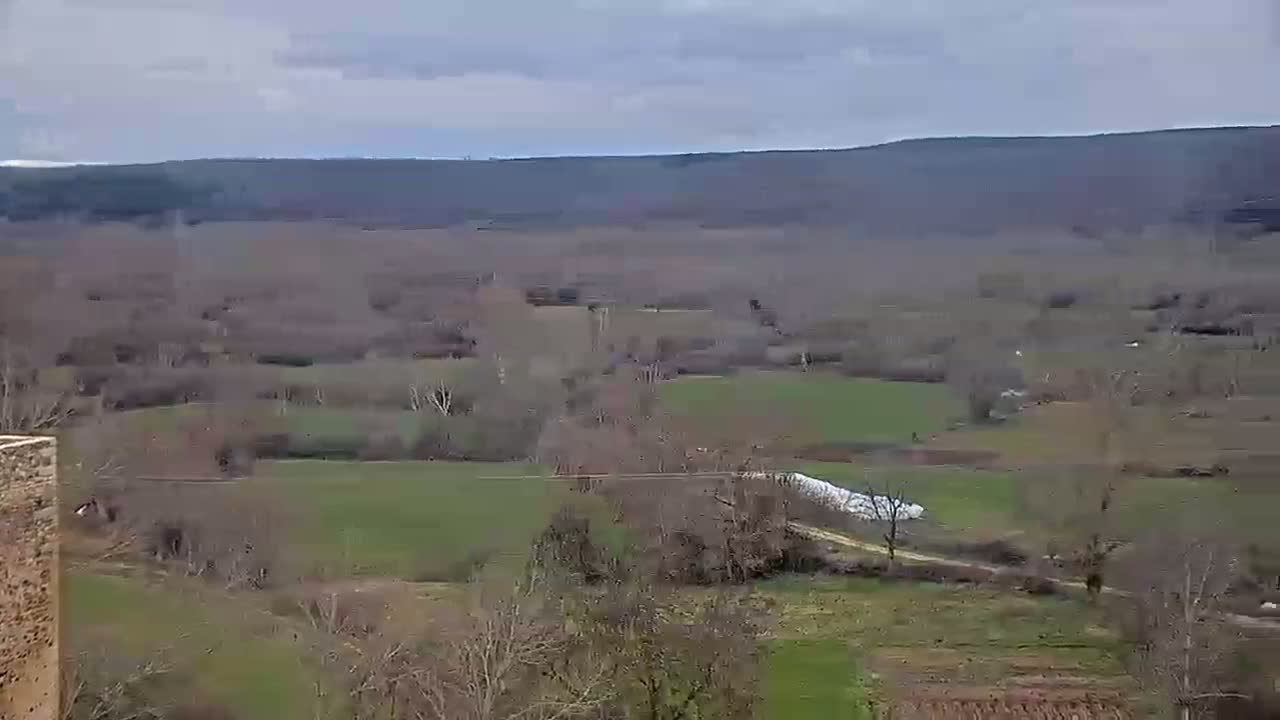 A high-angle view shows a vast, mostly treeless valley under an overcast sky, with patches of green fields and a dirt road winding through the landscape.