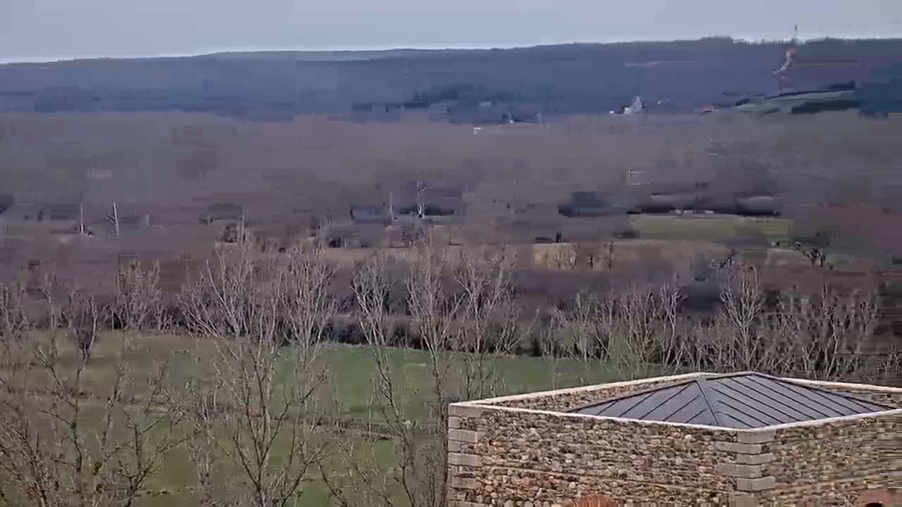 A stone structure with a metal roof sits in the foreground overlooking a vast, flat landscape of bare trees and fields under a cloudy sky.