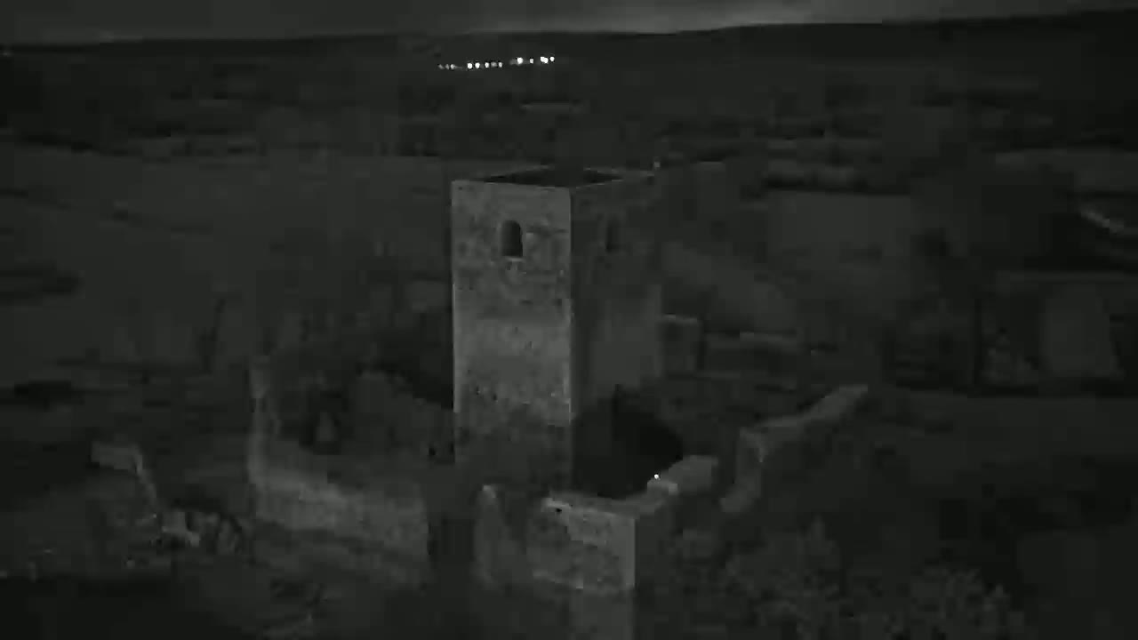 An aerial nighttime view shows the ruins of a stone structure, possibly a castle or tower, under a dark sky.