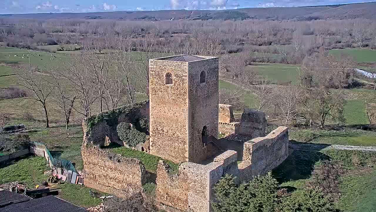 An aerial view shows the stone ruins of a castle or tower, partially overgrown with vegetation, situated in a rural landscape under a mostly sunny sky.