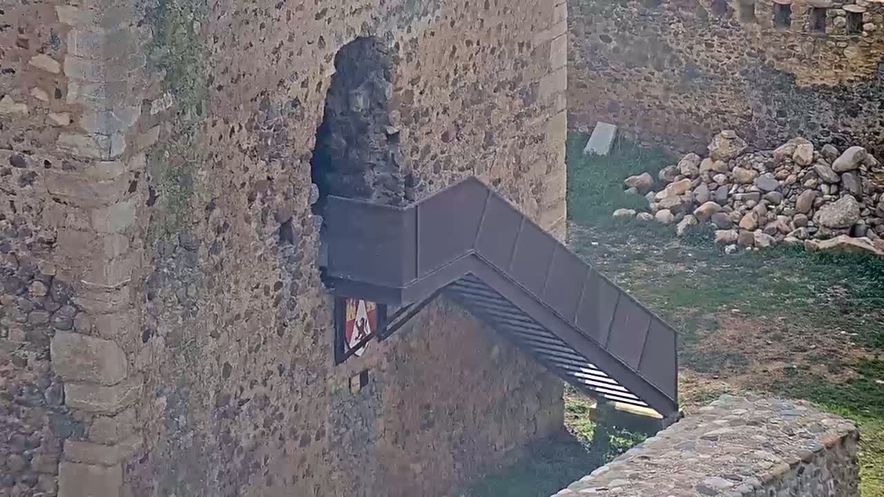 A modern metal staircase extends from a stone castle wall onto a grassy area with a pile of rocks nearby.