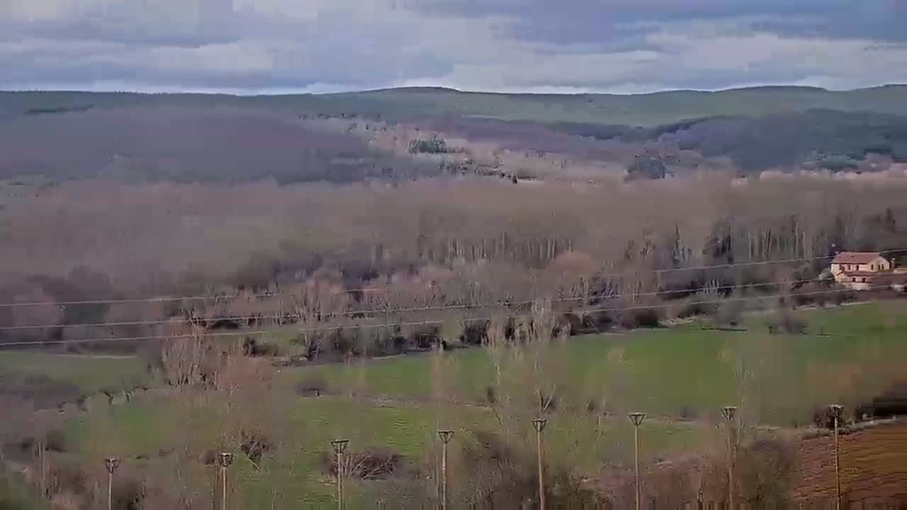 A view of a valley under a cloudy sky shows bare, winter trees, green fields, and a single building nestled among the trees.