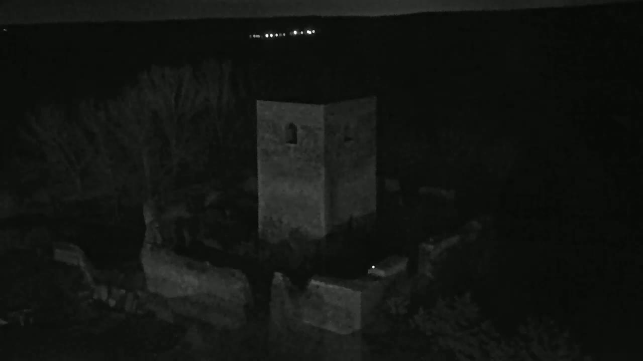 A nighttime, low-light image shows a square stone tower and surrounding structures, possibly ruins, under a dark sky with distant lights visible on the horizon.
