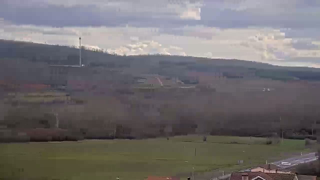 A mostly cloudy day reveals a landscape of rolling hills covered in muted green and brown vegetation, a distant communication tower, and a road running through the lower foreground.