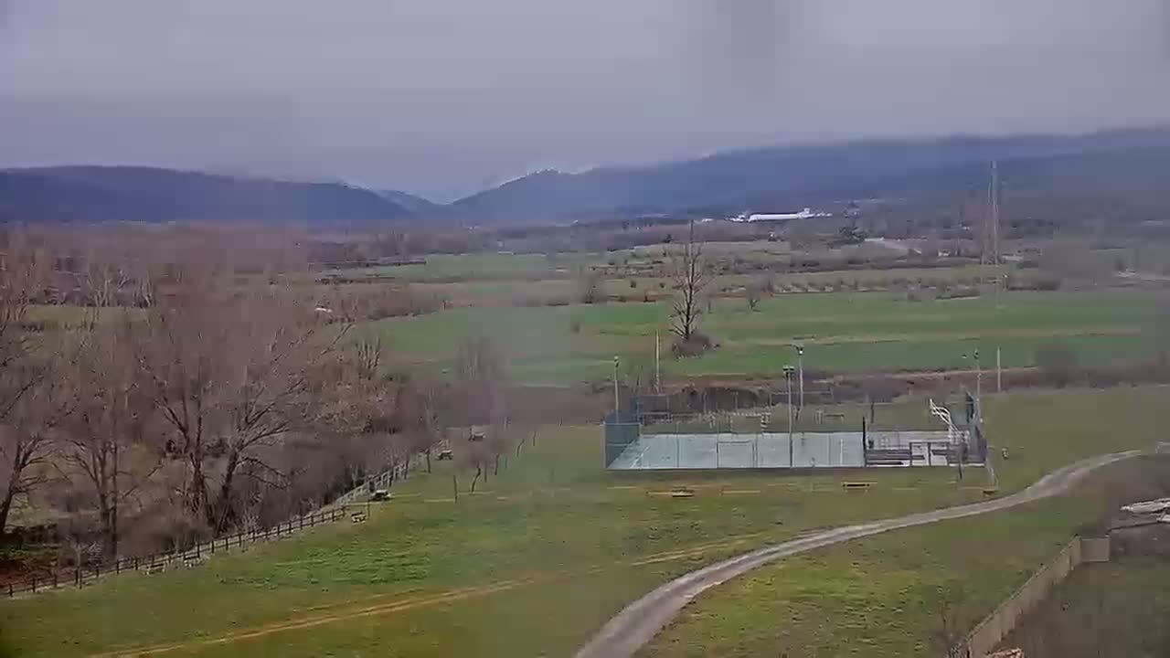 A panoramic view of a rural landscape under an overcast sky shows a valley with bare trees, green fields, and a tennis court near a dirt path.