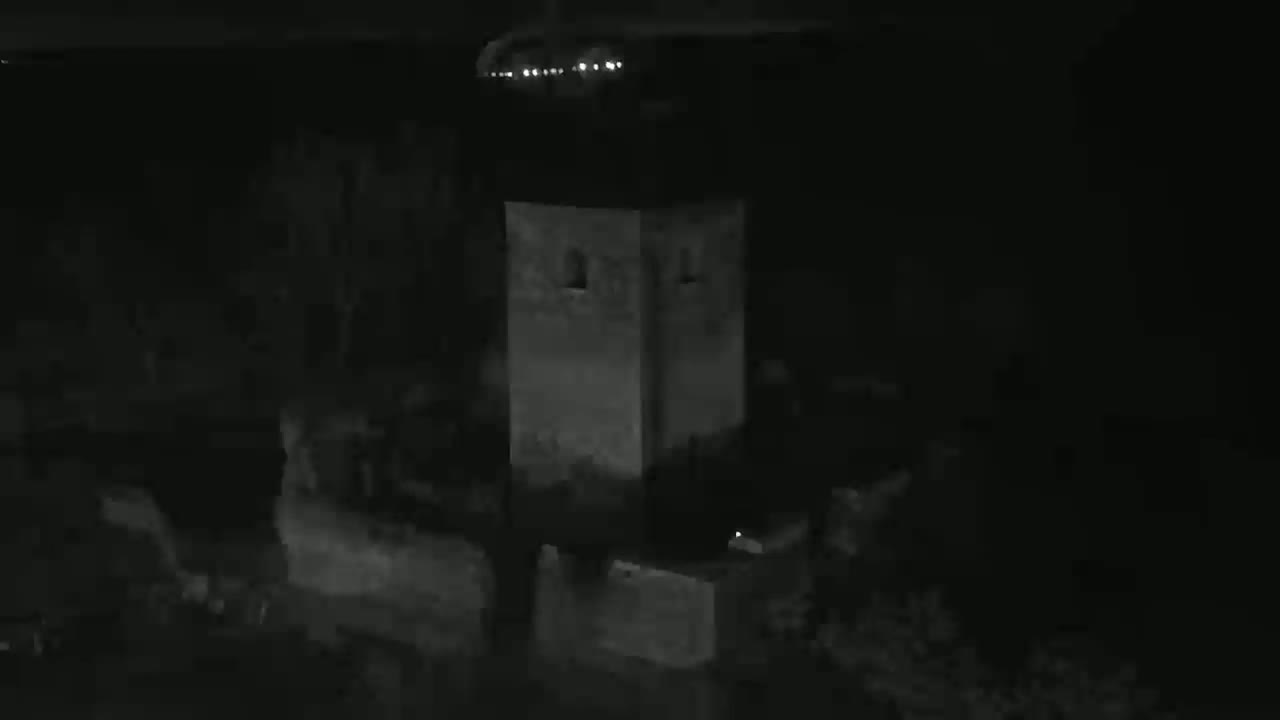 A dark, nighttime image shows a stone tower surrounded by shadowy vegetation under a dark sky.