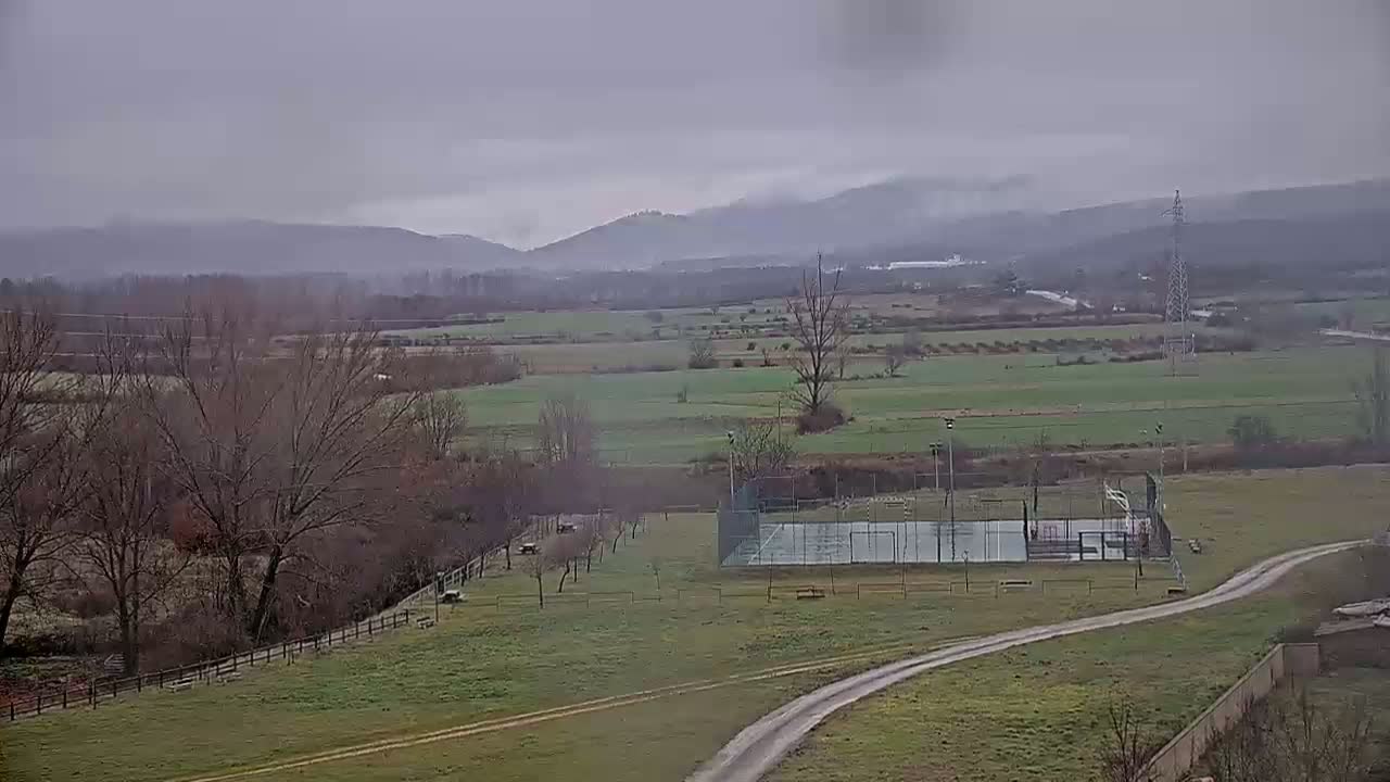 A sprawling, overcast landscape features a fenced-in sports court, bare winter trees, and rolling green hills in the distance.